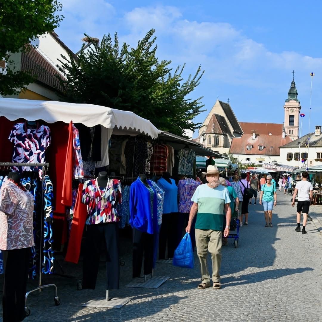 Ein geschäftiger Straßenmarkt mit verschiedenen Kleidungsständen unter Markisen, ein Mann mit einer blauen Tasche geht vorbei, und Menschen schlendern im Hintergrund.
