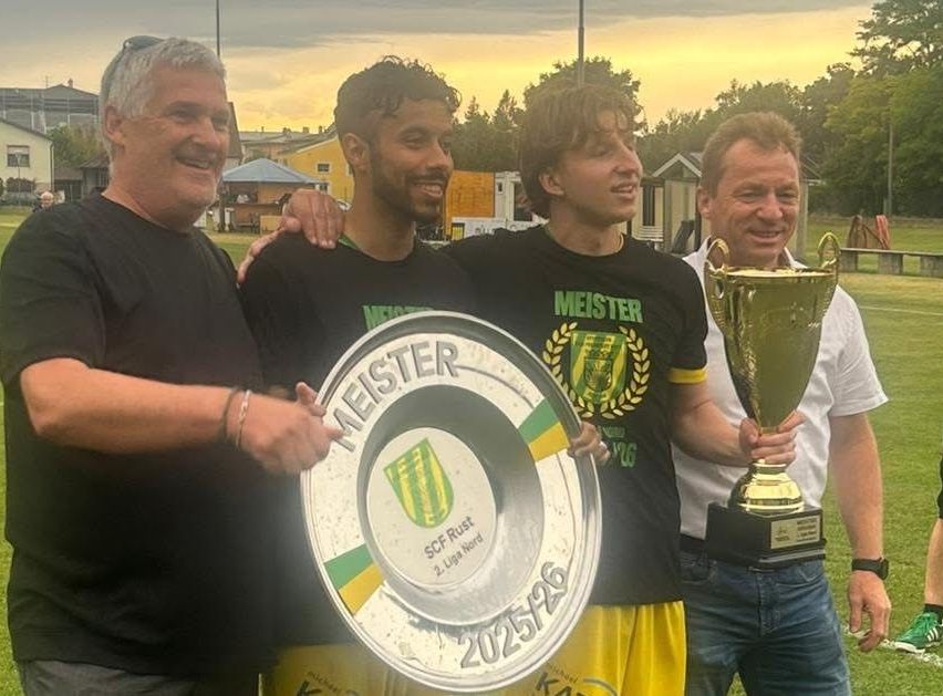 Vier Männer stehen lächelnd in einer Gruppe. Einer hält eine Trophäe und ein anderer eine Platte mit dem Wort Meister und einem Datum. Sie scheinen einen Sieg zu feiern.