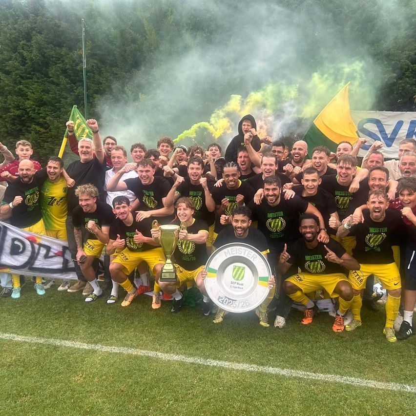 Eine Fußballmannschaft feiert mit einem Pokal und einem Schild auf dem Feld, mit Rauch und Flaggen im Hintergrund.