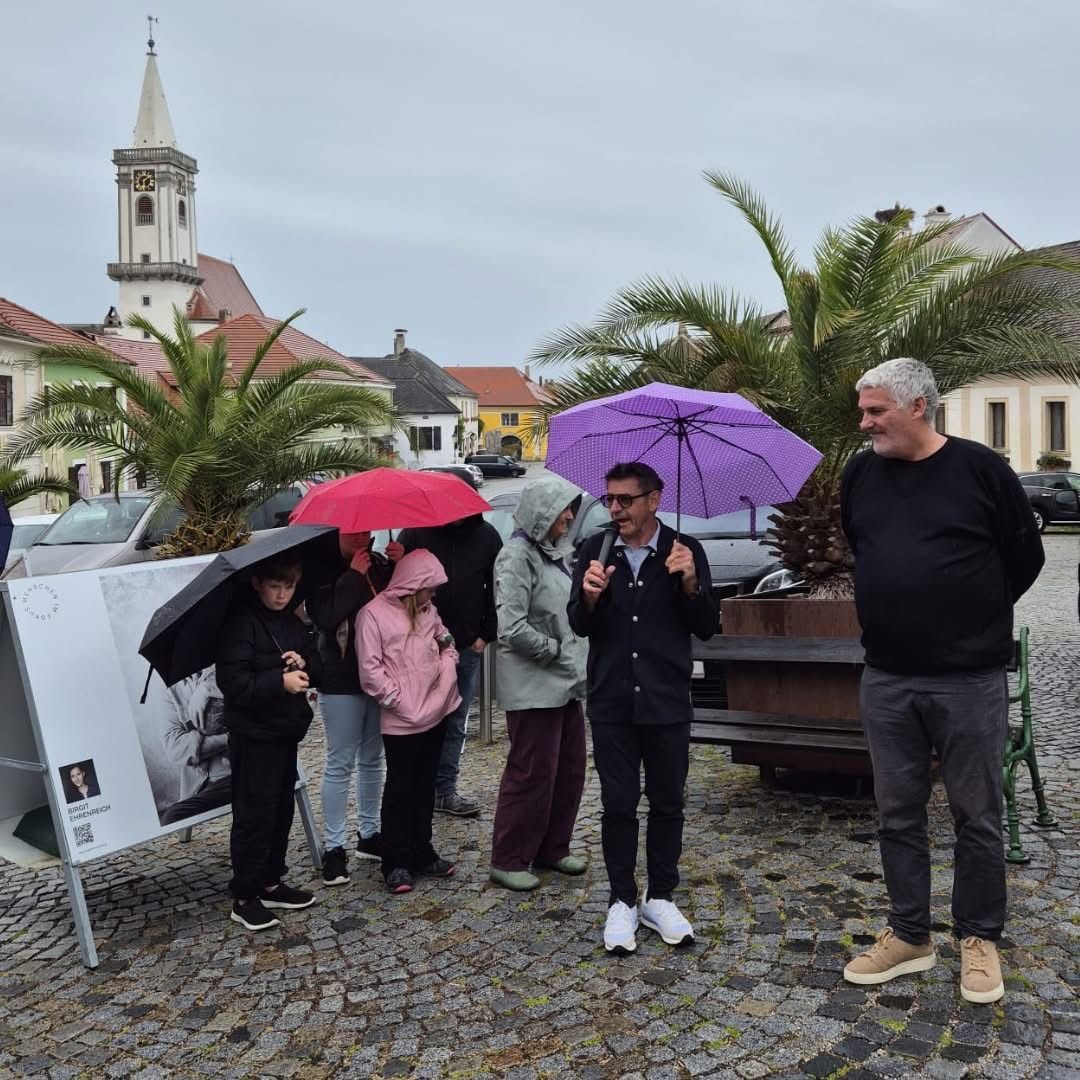Eine Gruppe von Menschen, einschließlich Kinder, steht unter Regenschirmen auf einem Stadtplatz. Sie stehen vor einer Kirche mit einem Turm, und ein Mann mit einem Mikrofon spricht zu ihnen.