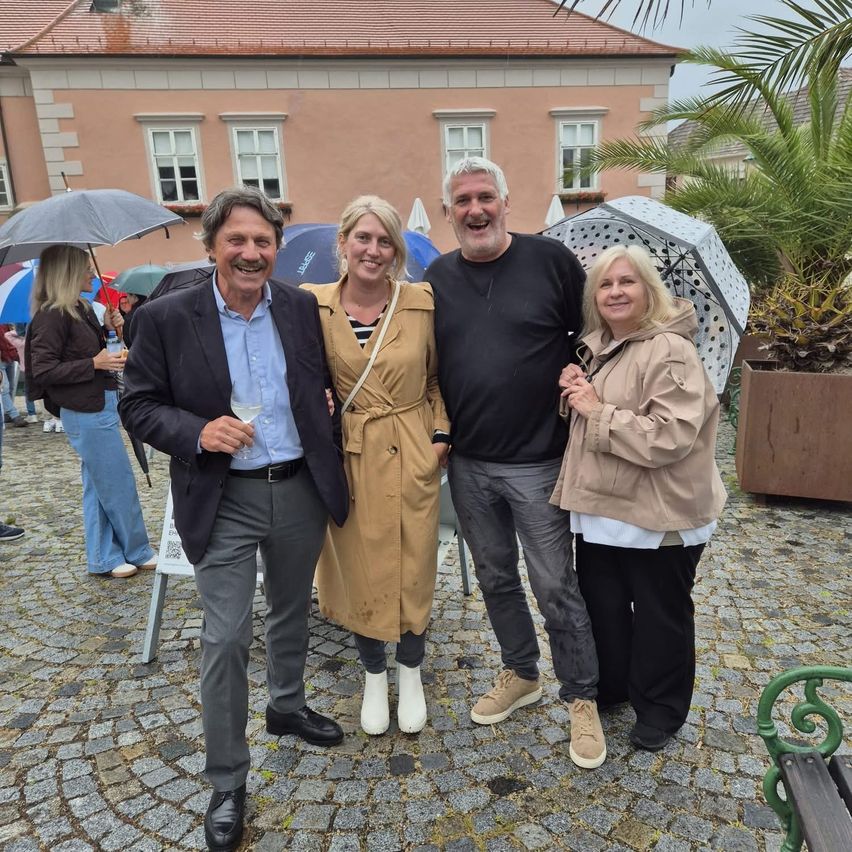 Eine Gruppe von Menschen steht draußen mit Regenschirmen. Sie lächeln und scheinen für ein Foto zu posieren. Das Wetter ist regnerisch.