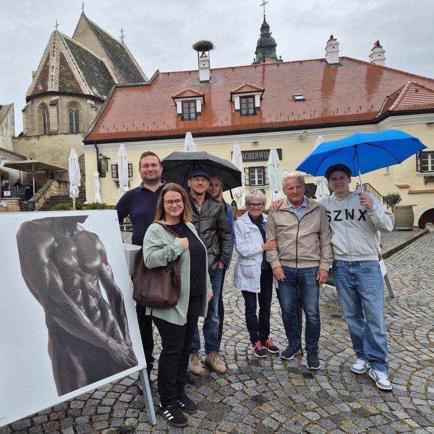 Eine Gruppe von Menschen posiert für ein Foto vor einem Gebäude mit roten Dächern. Sie halten Regenschirme und stehen neben einem großen Poster eines Mannes.