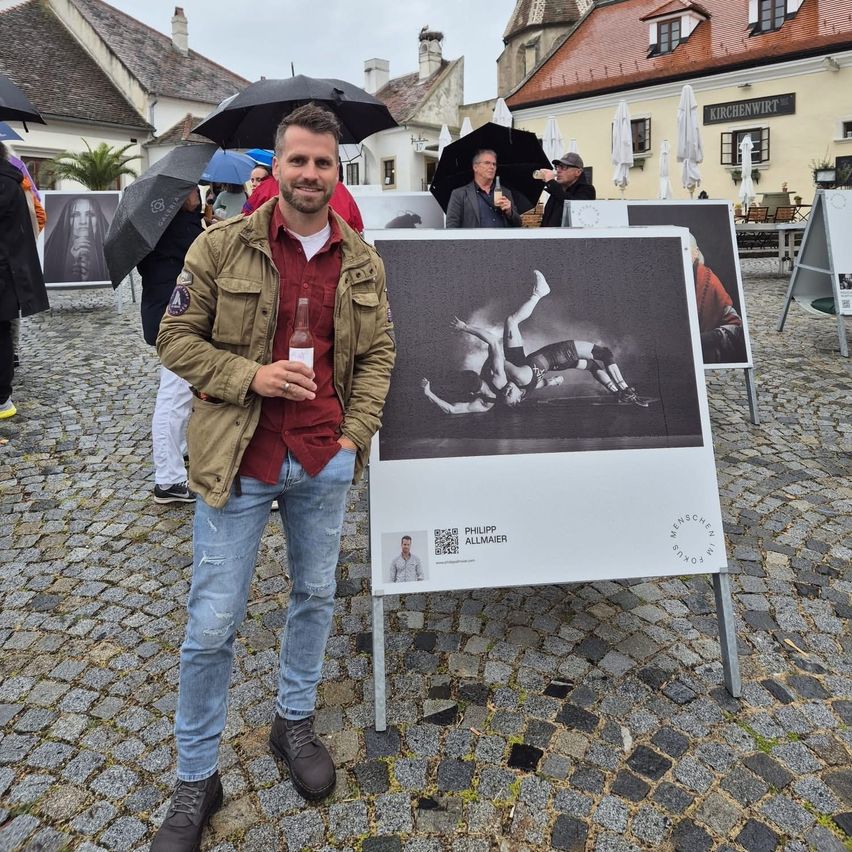 Ein Mann steht neben einer Fotoausstellung auf Kopfsteinpflaster und hält eine Flasche. Hinter ihm Leute mit Regenschirmen. Das Foto zeigt einen Mann in Bewegung, mit Text am unteren Rand.