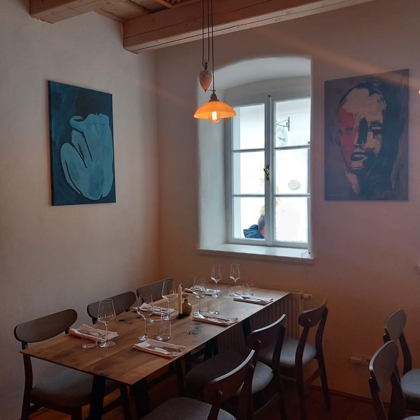 Ein leerer Essbereich mit einem holzigen Tisch für das Essen. Zwei abstrakte Gemälde hängen auf beiden Seiten eines Fensters. Außerhalb des Fensters ist eine Person in Blau zu sehen.