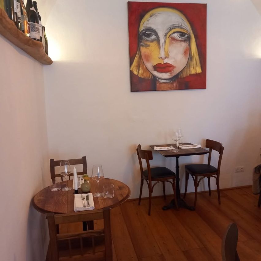 Ein kleiner Essbereich mit Holztischen, Stühlen und einem Gemälde an der Wand. Die Tische sind mit Gläsern, Servietten und Besteck gedeckt. Flaschen sind auf einem Regal.