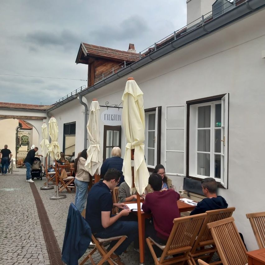Eine Gruppe von Menschen sitzt an einem Tisch vor einem Restaurant namens Freistein. Das Wetter ist bewölkt. Sonnenschirme und Stühle sind um die Tische herum aufgestellt.