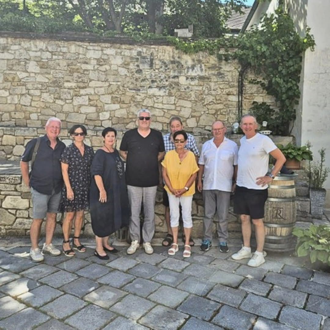 Eine Gruppe von Menschen steht vor einer Steinmauer mit Grünpflanzen und Fässern, wahrscheinlich auf einer Tour. Sie tragen Freizeitkleidung und teilweise Sonnenbrillen.