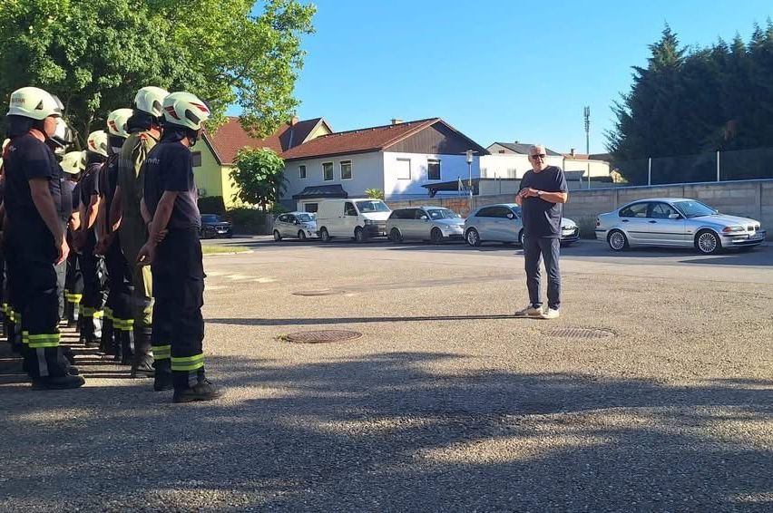 Ein Mann steht vor einer Reihe von Feuerwehrleuten auf einem Parkplatz mit mehreren geparkten Autos dahinter.