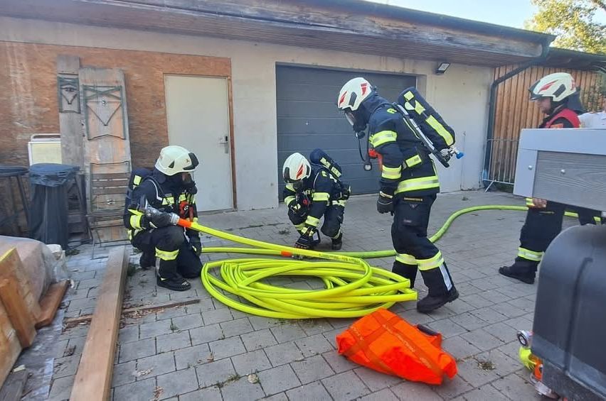 Feuerwehrleute verwenden einen gelben Schlauch auf einem Ziegelboden in der Nähe eines Gebäudes. Ein Feuerwehrmann hält den Schlauch, während andere daran arbeiten.