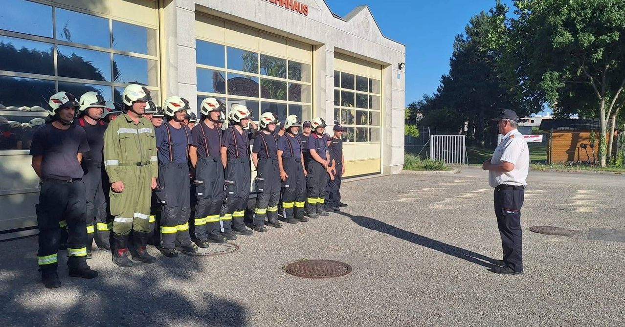 Eine Gruppe von Feuerwehrleuten steht in einer Reihe vor einem Feuerwehrhaus. Sie tragen Helme, Uniformen und Stiefel. Hinter ihnen spiegelt ein Gebäude mit Glasfenstern die Bäume draußen.