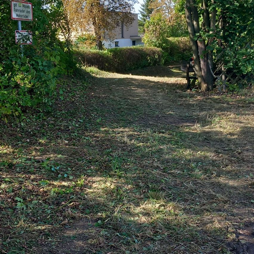 Ein schmaler Pfad mit einem Schild, das 'Fahrräder und Fußgänger nur' anzeigt, führt durch ein bewaldetes Gebiet mit einem Haus im Hintergrund.