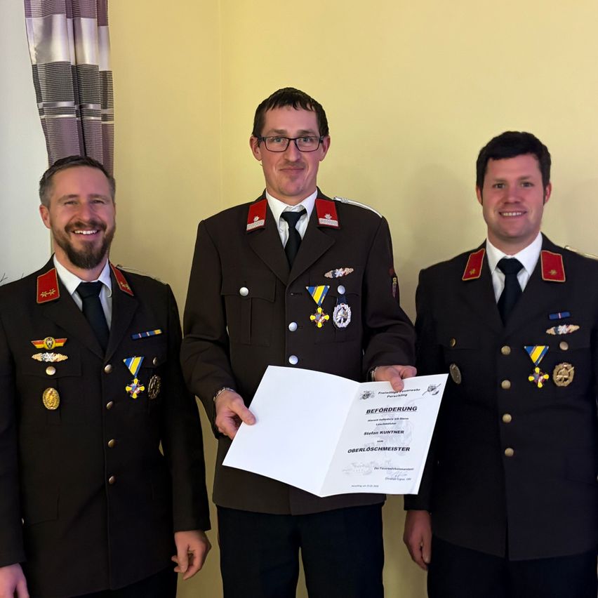 Drei Männer in Militäruniformen stehen und lächeln. Der Mann in der Mitte hält ein Zertifikat. Die Wand hinter ihnen ist gelb gestrichen.