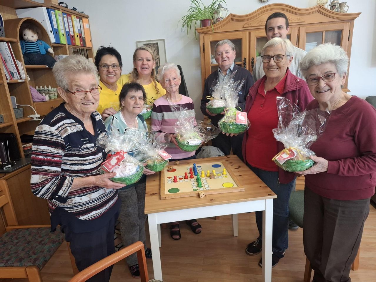A group of elderly individuals, both men and women, stand around a wooden table. They are all holding baskets with gifts. The table has a game board on it. Behind them is a cabinet with a potted plant and a framed picture.