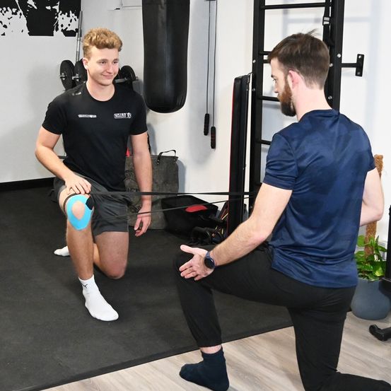 Ein Mann mit Kniestütze hockt sich und trainiert, während ein anderer Mann in einem Fitnessstudio zuschaut.