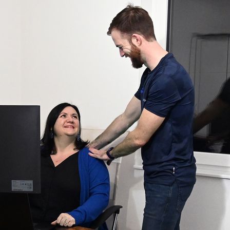 A man with a beard stands behind a woman, both smiling, as he gently touches her shoulder in an indoor setting with a computer monitor nearby.