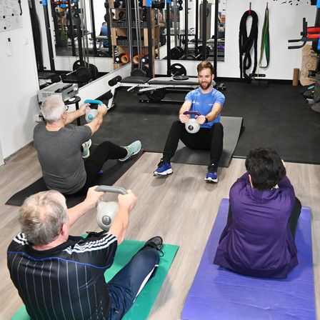 A group of people are exercising in a gym. Two men are sitting on a mat, one holding a kettlebell, and the other holding a weight. A man is standing in front of them.
