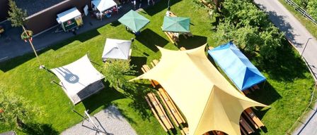 Ein Luftbild eines Parks zeigt ein großes gelbes Zelt, mehrere kleinere Zelte und ein Gebäude mit flachem Dach. Der Park hat grünen Rasen, Bäume und einen Spielplatz.