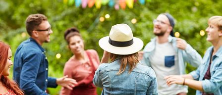 Eine Gruppe von Freunden plaudert und lacht in einem Garten mit dekorativen Lichtern. Eine Frau mit einem weißen Hut dreht sich um, um mit ihnen zu sprechen.