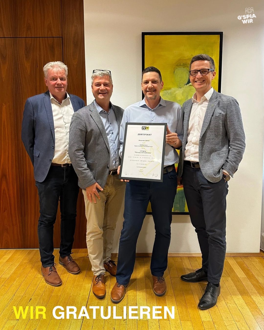 Vier Männer stehen lächelnd zusammen in einem Raum, einer hält ein Zertifikat. Sie tragen formelle Kleidung und stehen vor einer Holzwand mit einem farbenfrohen Gemälde.