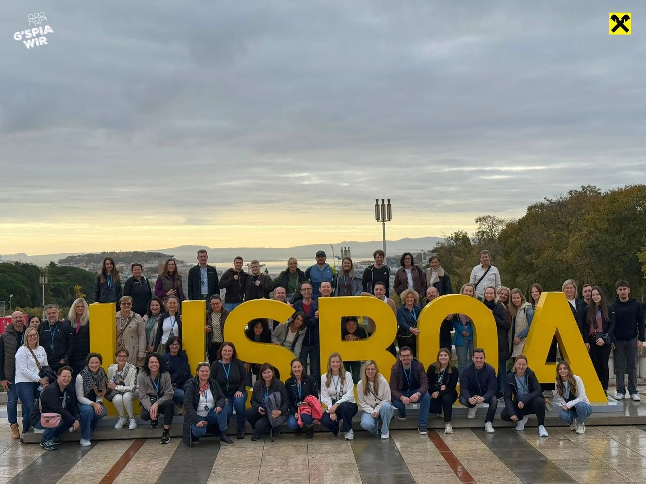 Eine Gruppe von Menschen posiert für ein Foto vor einem großen gelben Schild, auf dem Lissabon steht. Sie stehen auf einem gefliesten Boden, einige sitzen.