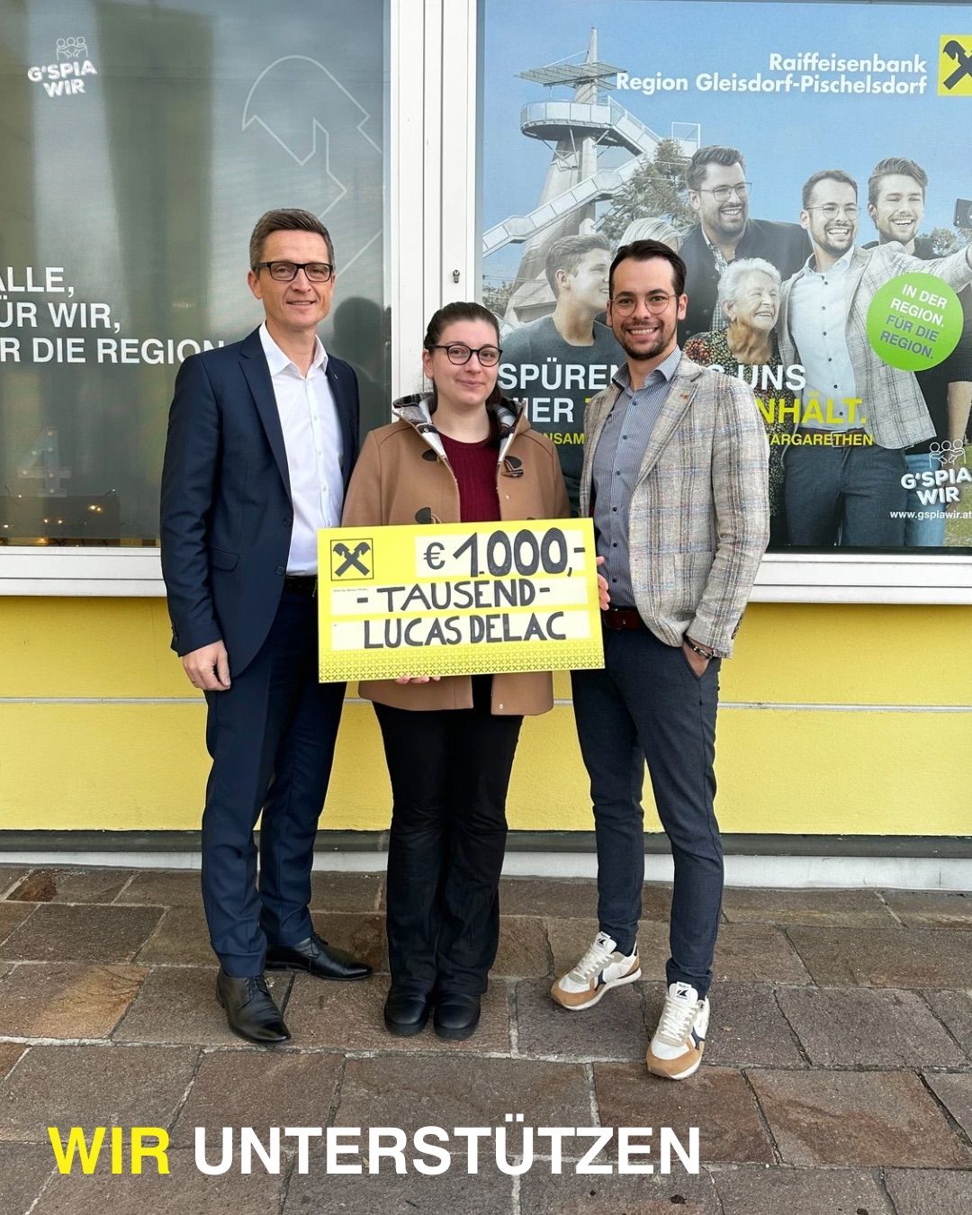 Drei Personen stehen auf einem Bürgersteig. Eine Frau hält ein gelbes Schild mit dem Text '1000 Euro für Lucas Delac'. Dahinter befindet sich ein Plakat mit mehreren Personen.