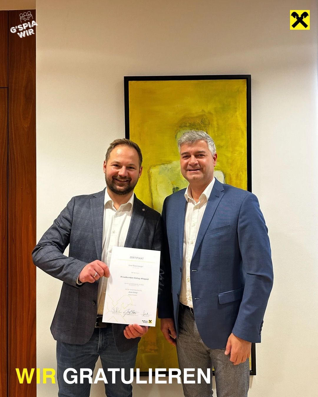 Zwei Männer stehen vor einem gelben Gemälde. Der Mann auf der linken Seite hält ein Zertifikat und lächelt. Der Mann auf der rechten Seite trägt einen blauen Anzug und lächelt.