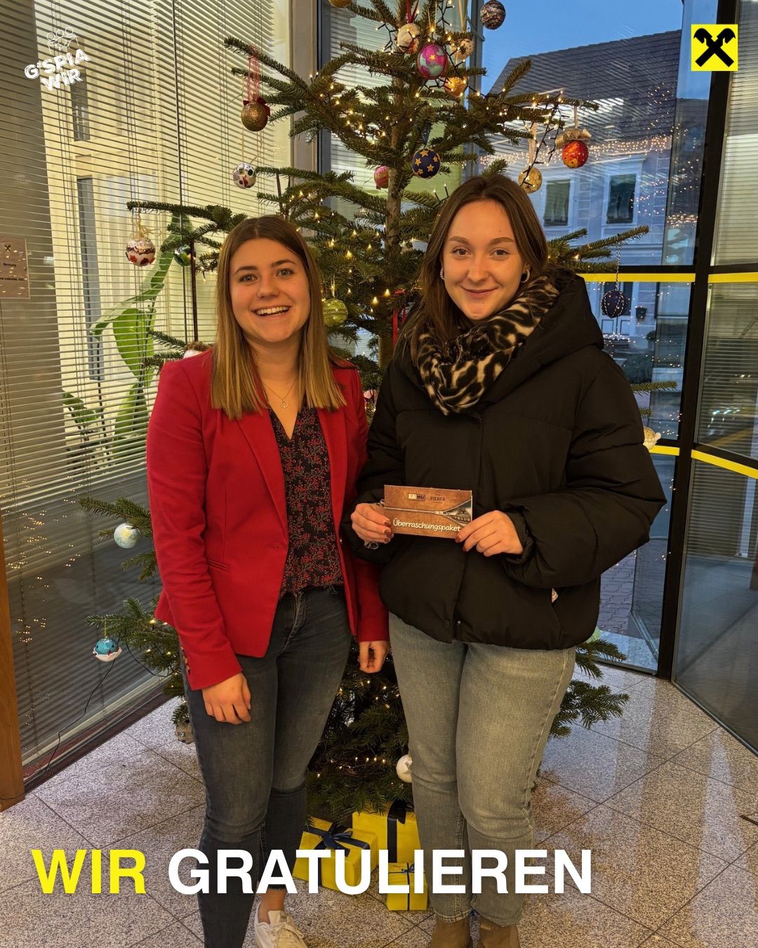 Zwei Frauen stehen vor einem geschmückten Weihnachtsbaum und halten eine Geschenkbox. Die Frau auf der linken Seite trägt einen roten Blazer und die Frau auf der rechten Seite trägt eine schwarze Daunenjacke. Sie lächeln beide.