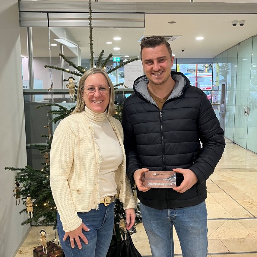 Ein Mann und eine Frau lächeln vor einem Weihnachtsbaum. Der Mann hält eine Schachtel und sie scheinen beide zu feiern.