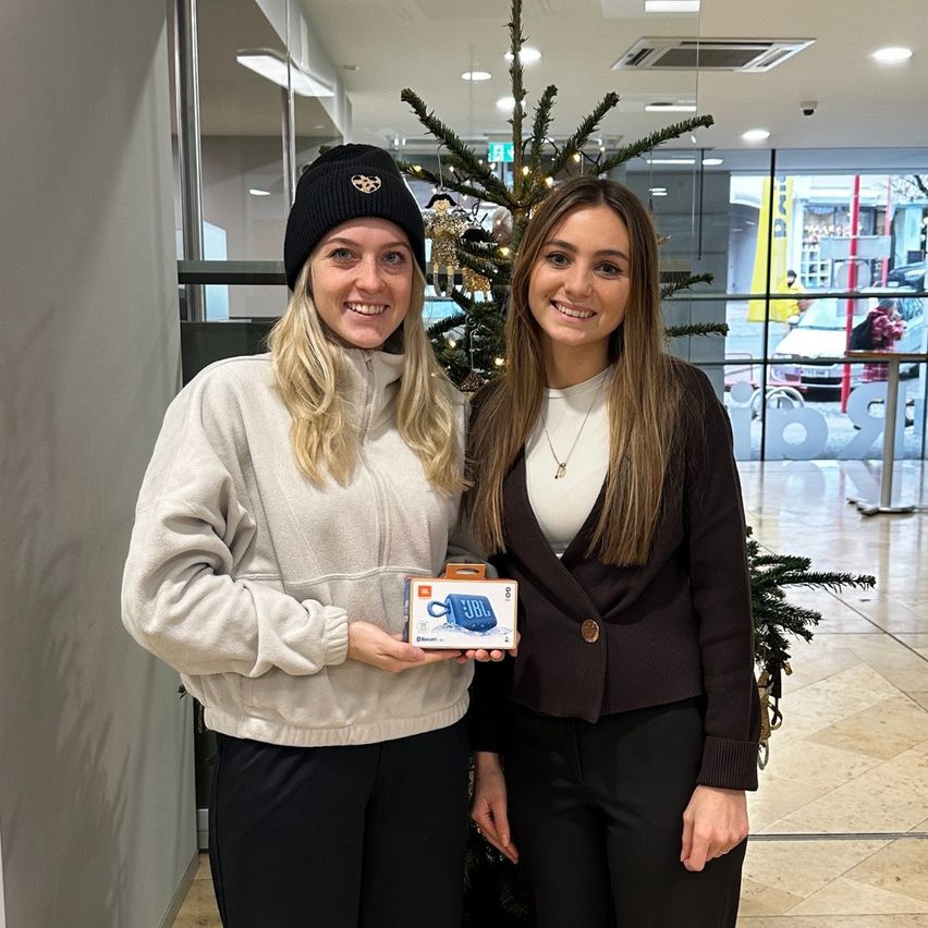 Zwei Frauen stehen neben einem Weihnachtsbaum und lächeln. Eine hält eine JBL-Box für einen tragbaren Lautsprecher. Sie befinden sich in einem Einkaufszentrum in der Nähe einer Glaswand, die Autos und einen gelben Banner zeigt.