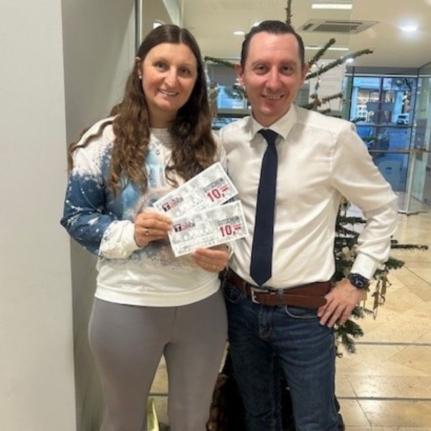 Eine Frau und ein Mann stehen neben einem Weihnachtsbaum und lächeln. Die Frau hält mehrere Tickets in der Hand, von denen eines sichtbar ist und die Nummer 10 zeigt.