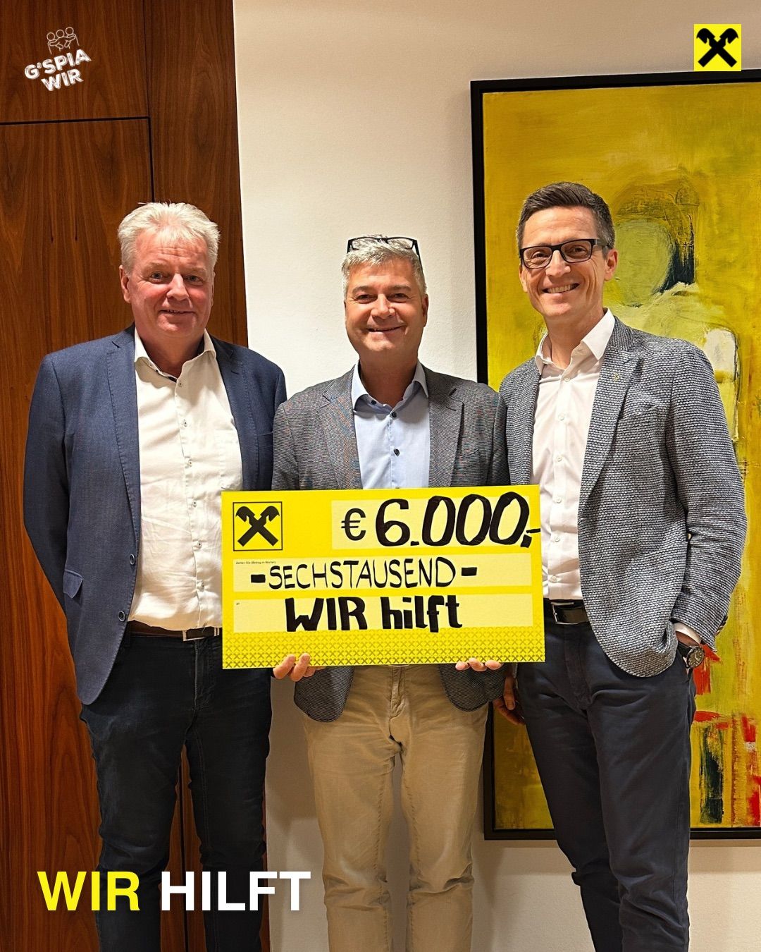 Three men stand close together in an indoor setting, smiling for the camera. The man in the middle holds a yellow sign that reads '€ 6,000.- SEKHSTAUSEND WIR hilft'. They are in front of a bright yellow and white abstract painting.