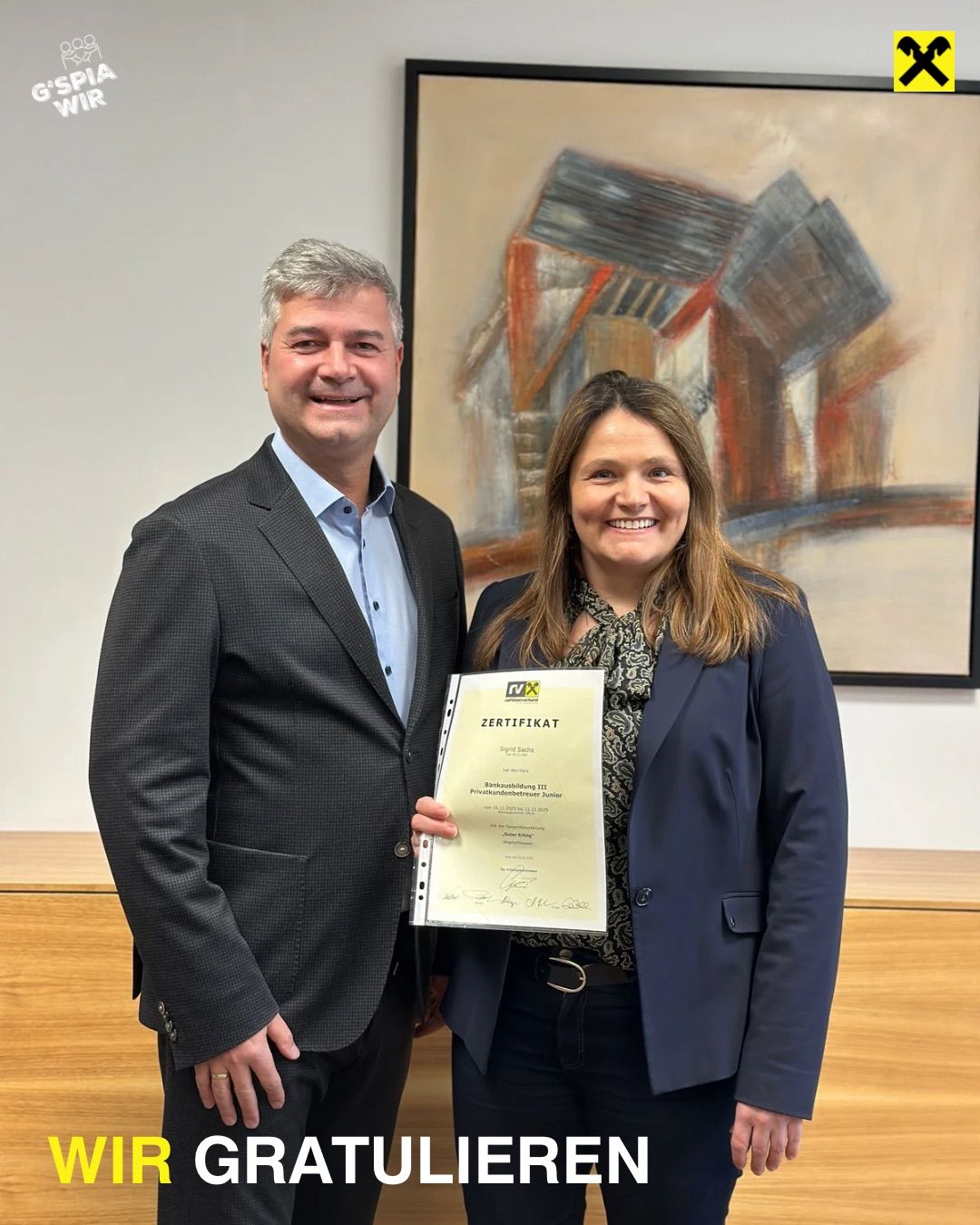 Ein Mann und eine Frau stehen lächelnd in einem Büro. Die Frau hält ein Zertifikat mit gelber Umschlag. Der Mann trägt einen Anzug, und die Frau trägt einen marineblauen Blazer. Hinter ihnen hängt ein großes Gemälde an der Wand.