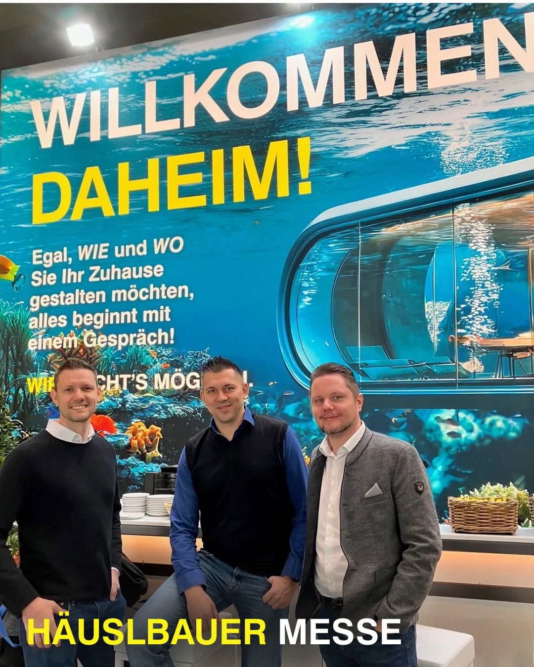 Drei Männer stehen lächelnd vor einem großen Wandbild. Das Wandbild zeigt eine Unterwasserszene mit einer Glasblase und buntem Meeresleben. Über der Szene steht auf Deutsch 'Willkommen Daheim!'.