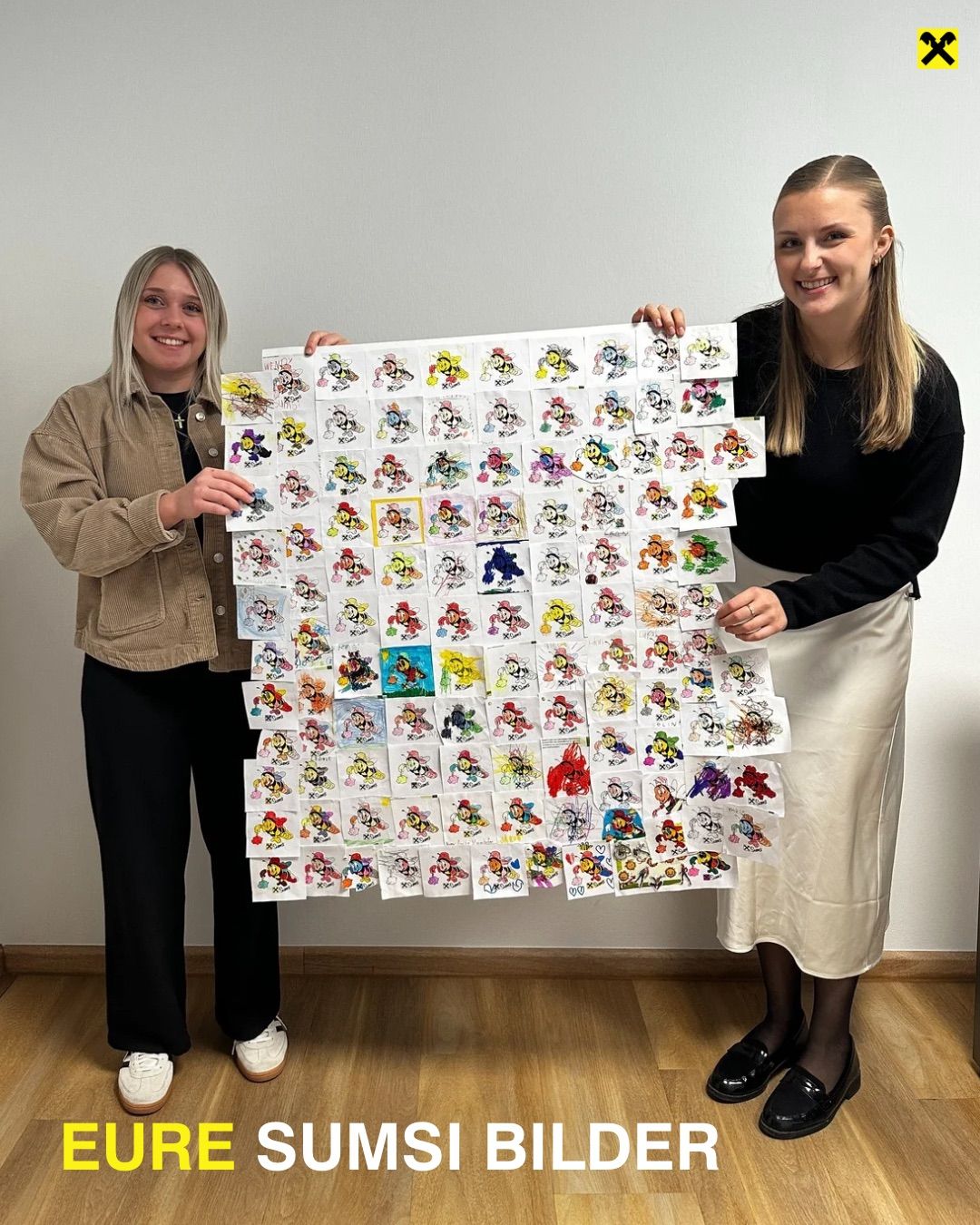 Two women are standing and holding a large collage of colorful cartoon character drawings. They are smiling and appear to be posing for a photo. They are in a room with a wooden floor.