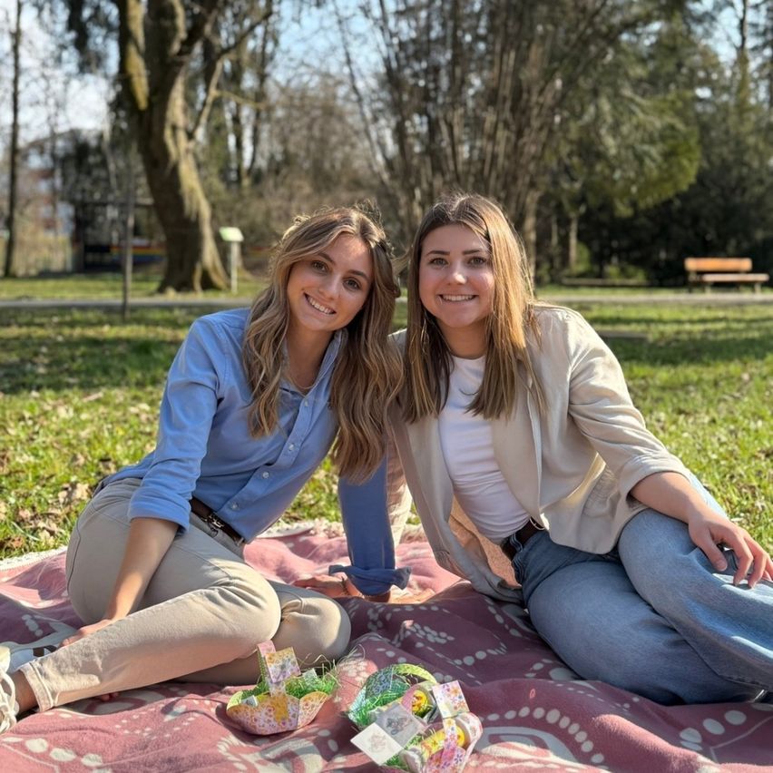 Zwei Frauen sitzen auf einer Picknickdecke im Park und lächeln für ein Foto. Eine hat blonde Haare und trägt ein blaues Shirt, während die andere eine beige Jacke trägt. Sie sind von Osterkörben und Bäumen umgeben.