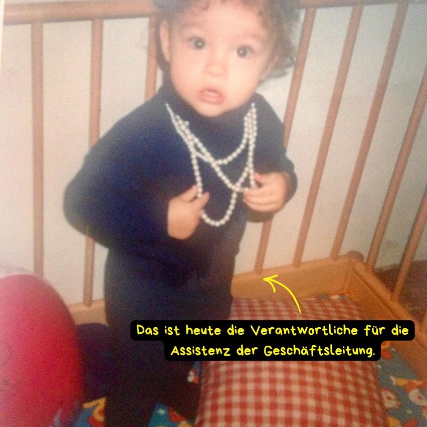 A toddler in a crib wearing pearl necklaces looks serious. There is a red ball and a yellow arrow pointing at the child.