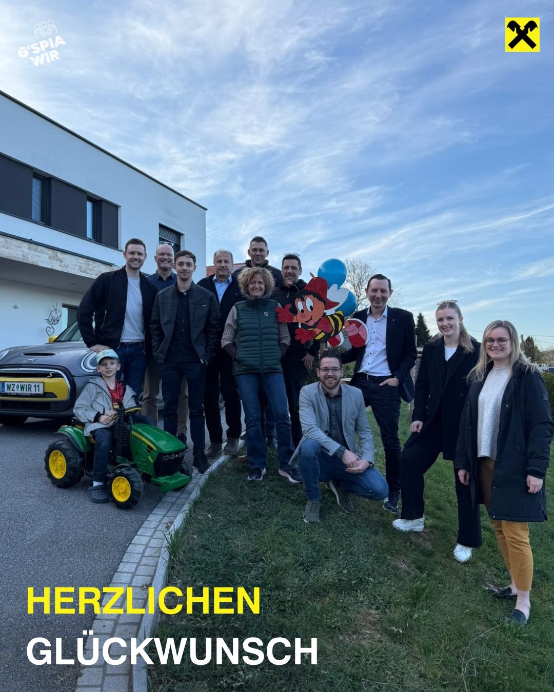 A group of people stand in front of a building, smiling. A child rides a toy tractor, and a car is parked. A sign reads Herzlichen.