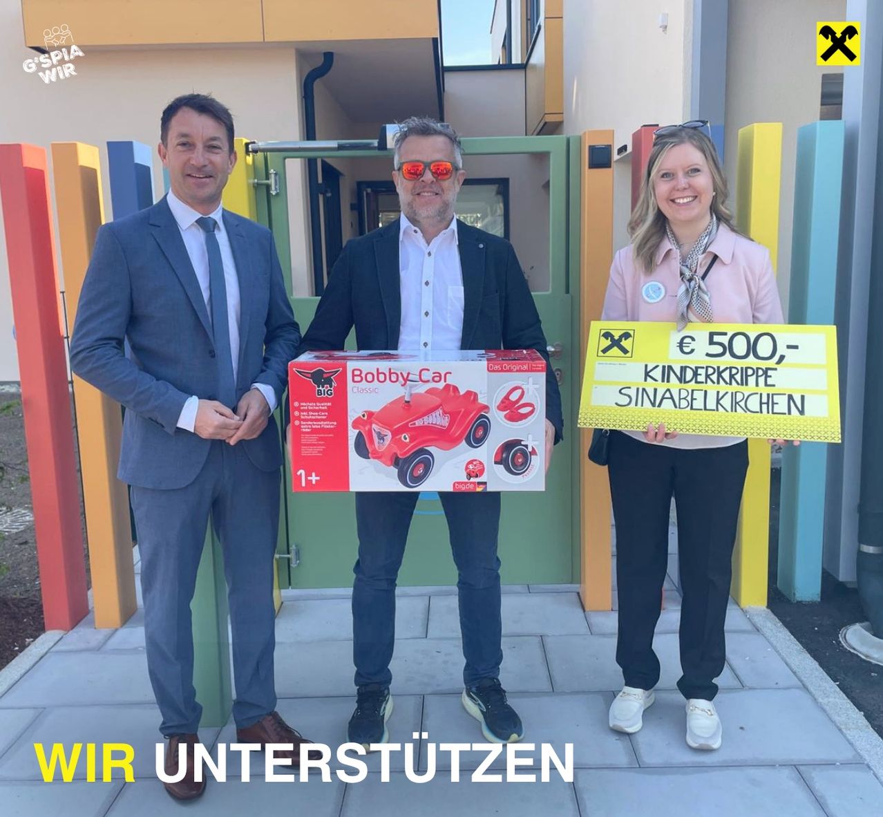 Drei Personen stehen vor einer gelben Tafel mit der Aufschrift 500€ für Kinderkrippe Sinabelkirchen. Ein Mann hält eine Schachtel mit der Aufschrift Bobby Car.