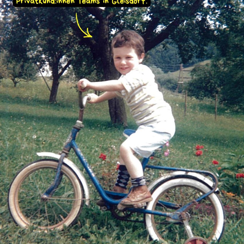 Ein kleiner Junge fährt ein blaues Fahrrad mit Stützrädern auf einer Wiese. Der Junge trägt ein gestreiftes Hemd und braune Schuhe. Hinter ihm steht ein Baum und ein verschwommener Hintergrund.