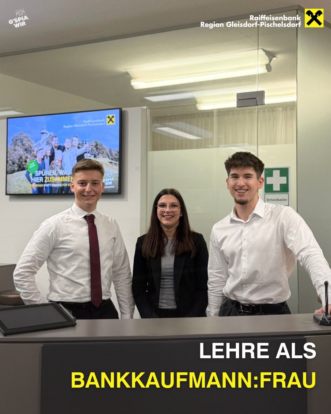 Drei Personen stehen lächelnd vor einem Empfangstresen für ein Foto. Eine Frau trägt eine Brille und einen schwarzen Blazer. Zwei Männer tragen weiße Hemden und Krawatten. Ein Bildschirm zeigt ein Plakat an.