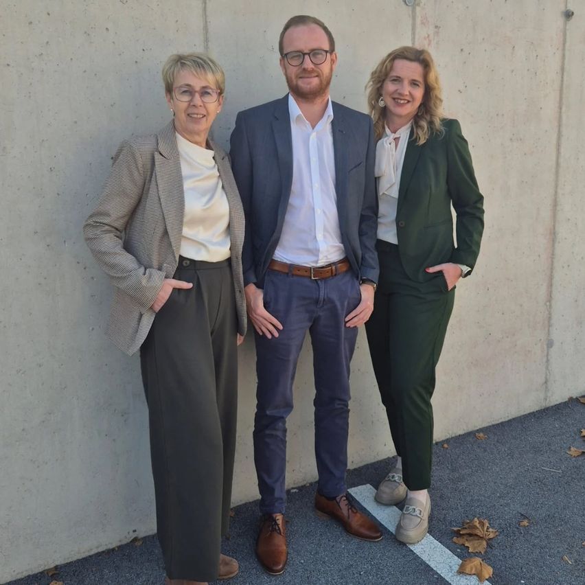 Three people stand next to each other, smiling, against a concrete wall. A woman in glasses and a blazer, a man in a suit, and another woman in a green suit.