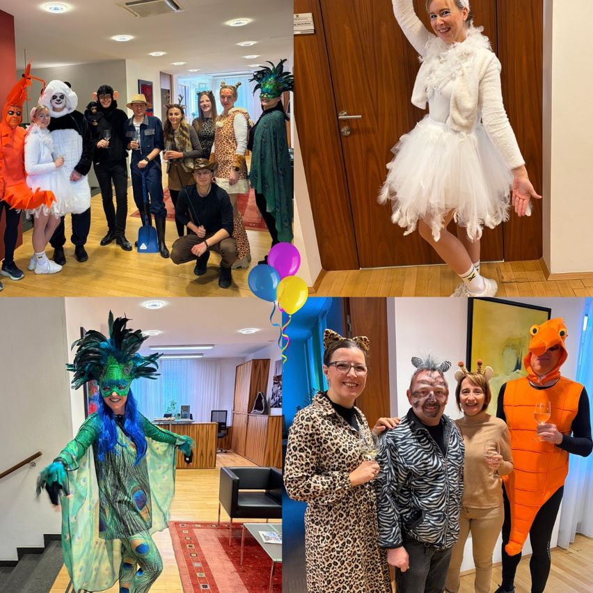 Eine Gruppe von Menschen in Tierkostümen und bunten Outfits posiert für ein Foto in einem Raum. Eine Frau trägt ein weißes, flauschiges Kostüm, während andere Kostüme wie Giraffe, Zebra und orange Monster tragen. Sie halten Gläser mit Wein.