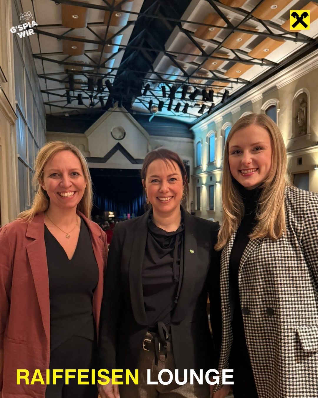 Drei Frauen stehen lächelnd in einem Saal. Die Frau auf der linken Seite trägt eine hellrosa Jacke. Die Frau in der Mitte hat eine schwarze Jacke. Die Frau auf der rechten Seite trägt einen schwarz-weiß karierten Mantel.