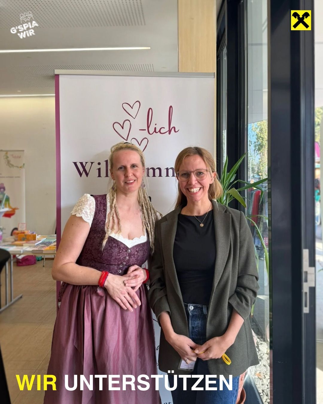 Zwei Frauen stehen lächelnd vor einem Schild mit dem Wort 'Wilmnn' und Herzen darauf. Die Frau auf der linken Seite trägt ein traditionelles Kleid, während die andere eine Jacke trägt.