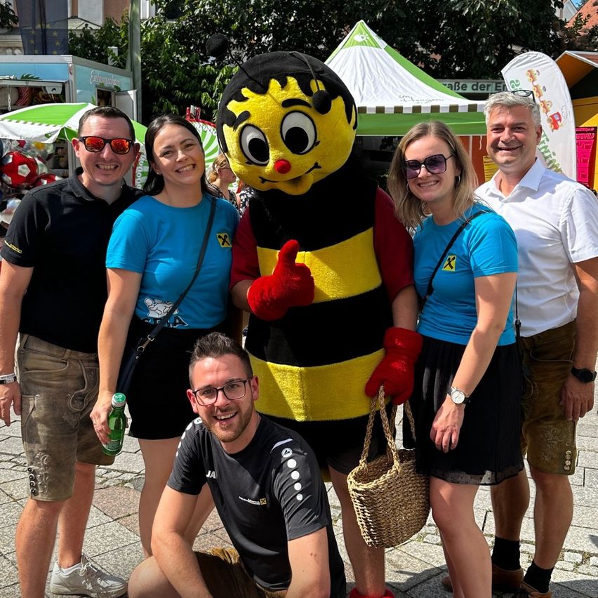 Fünf Personen posieren für ein Foto mit einem Bienen-Maskottchen in einer lebhaften Outdoor-Umgebung, möglicherweise bei einem Festival. Das Maskottchen ist in einem Bienenkostüm gekleidet und gibt einen Daumen hoch.