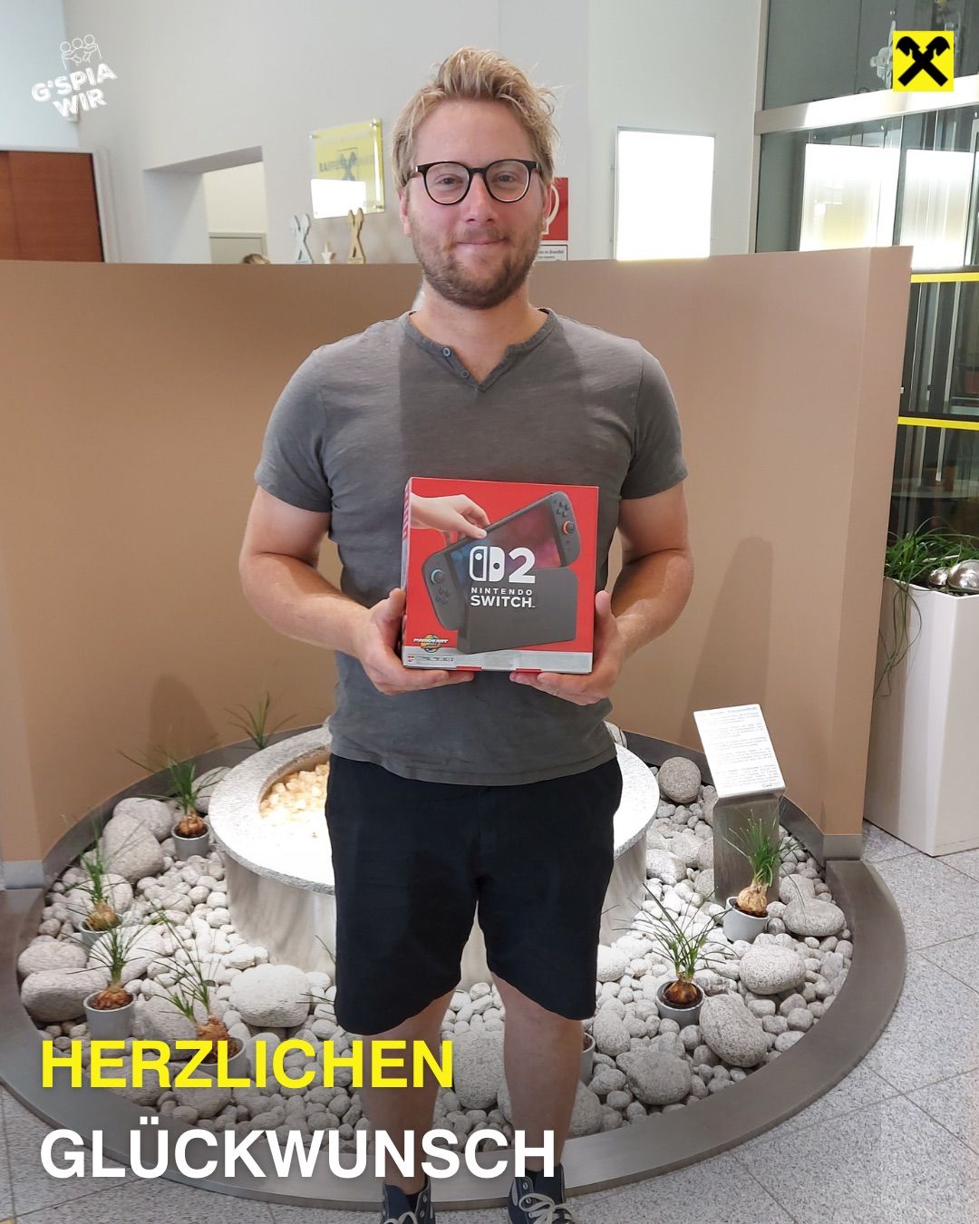 Ein Mann in einem grauen T-Shirt und schwarzen Shorts hält eine Nintendo Switch 2-Box vor einem dekorativen Steingarten.