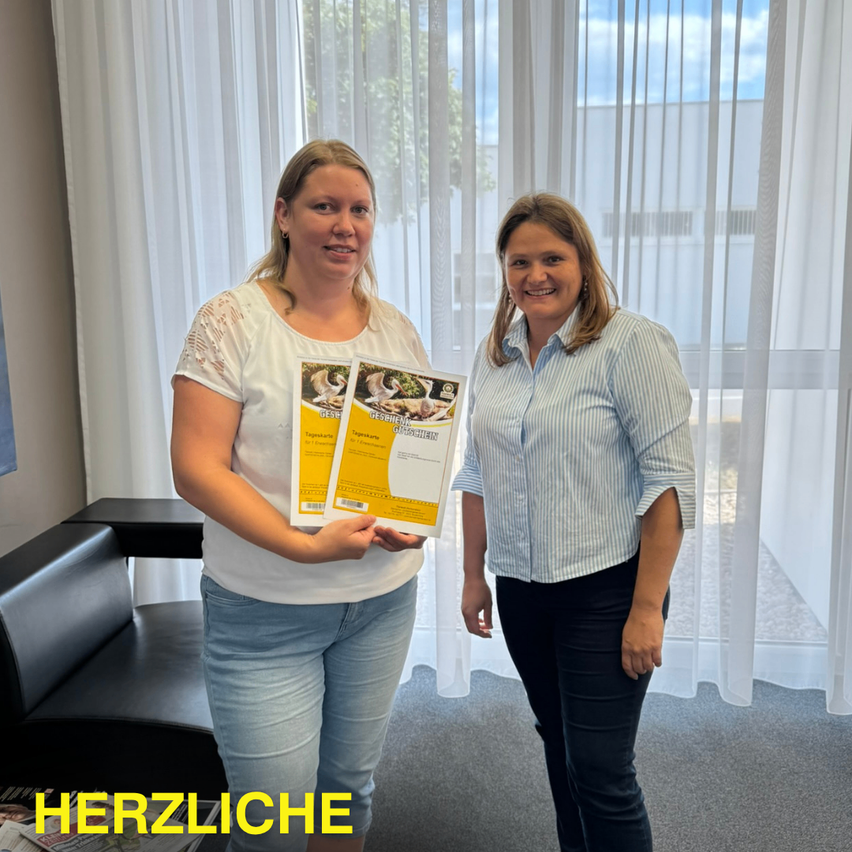 Zwei Frauen stehen nebeneinander in einem Büro, eine hält ein gelbes Heft mit einem Bild eines Babys auf dem Cover. Sie lächeln und stehen vor einem weißen Vorhang.