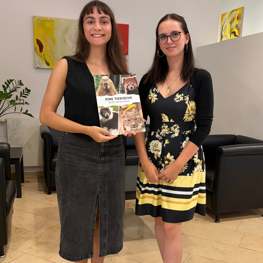 Zwei Frauen stehen in einem Raum und halten ein Buch mit dem Titel 'Eine Tierische Reise Um Die Welt'. Die Frau auf der linken Seite trägt ein schwarzes Kleid, während die Frau auf der rechten Seite ein gelbes Blumenkleid trägt. Hinter ihnen stehen schwarze Stühle und eine weiße Wand mit Gemälden.
