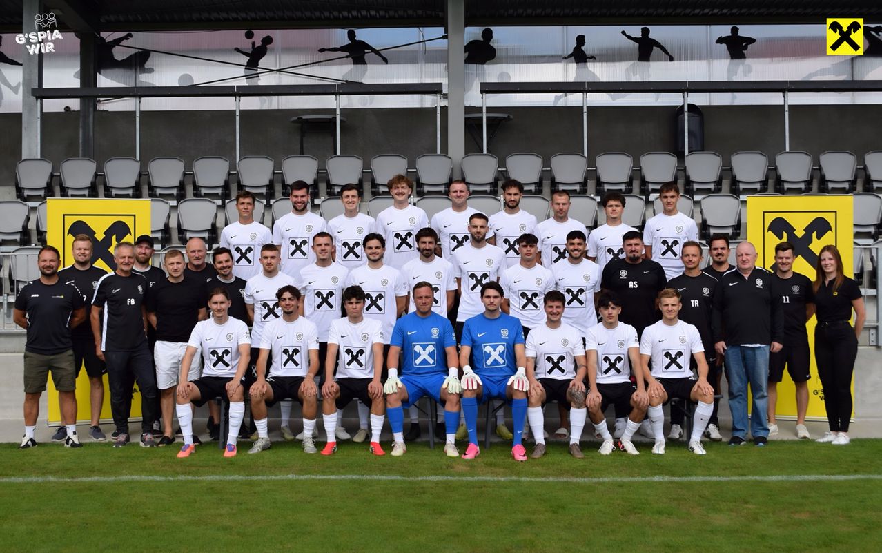 A soccer team poses for a group photo on a field, wearing white jerseys with black 'X' designs. The goalkeeper in blue stands out. Empty stadium chairs are in the background.