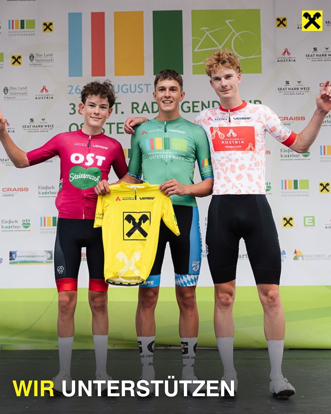 Drei junge Radfahrer stehen auf einem Podium und lächeln. Der auf der linken Seite trägt ein rosa Trikot mit 'OST' und 'Steiermark'. Der in der Mitte hält ein gelbes Trikot mit einem schwarzen X. Der auf der rechten Seite trägt ein weißes Trikot mit 'Cycling Austria'. Sie stehen auf einer Bühne mit einem farbenfrohen Hintergrund.